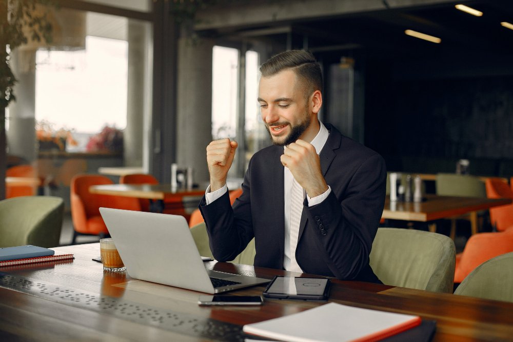 Businessman in suit celebrating success while working on laptop in modern office café.