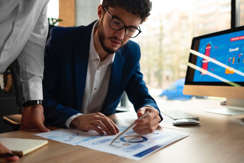 A businessman in a blue suit and glasses sits at a desk, using a stylus to point at data on a printed financial report.