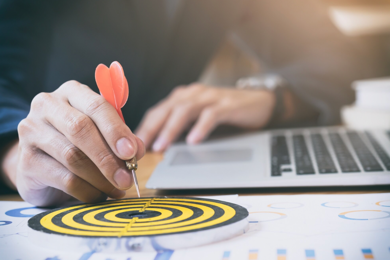 Hand placing dart on bullseye target beside laptop, symbolising business goal achievement.