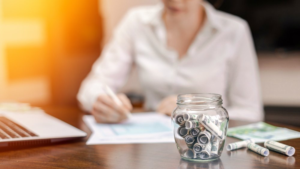 A clear glass jar filled with rolled-up US dollar bills sits prominently on a wooden desk.