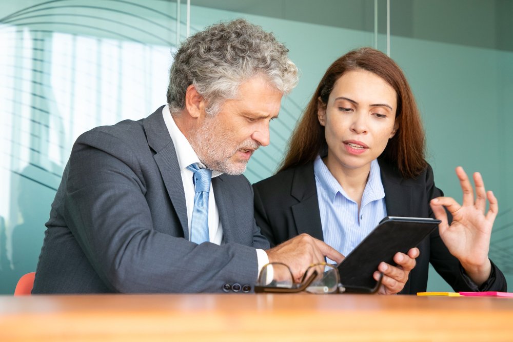 An experienced businessman and a female colleague are engaged in a serious discussion while looking at a tablet together.